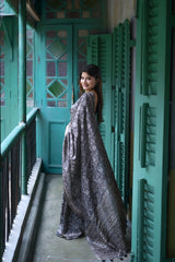 A woman models an elegant Black Tussar Silk Saree. The saree features dense all-over floral embroidery/weaving in a shimmering metallic silver thread. She wears a black sheer-sleeve blouse and heavy oxidized silver jewelry, including a large ring and stacked bangles, posing in a doorway framed by vibrant teal shutters.
