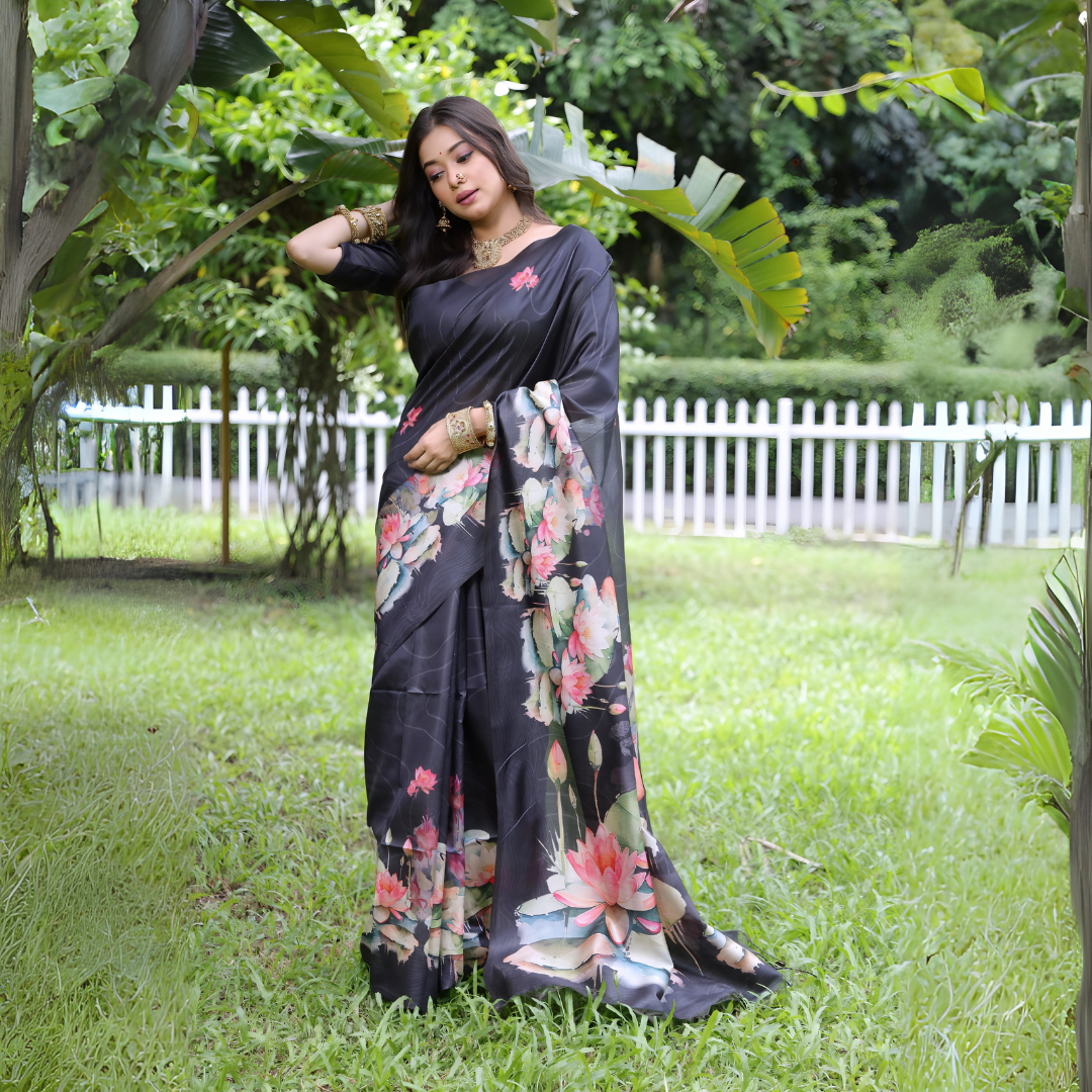 A woman models a stunning Deep Charcoal Black Soft Tussar Silk Saree. The saree features a large-scale digital print of colorful floral and lotus motifs in pink, coral, white, and green on the pallu and lower drape. She wears a matching elbow-sleeve black blouse and heavy gold Kundan jewelry, posing outdoors in a garden setting.