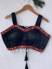 Black ethnic blouse featuring vibrant mirror work and multicolor embroidered border in red, green, blue, and yellow, adorned with pearl tassels and a tie-up at the back, displayed on a wooden hanger against a textured off-white wall with peacock feathers for a festive and traditional look.