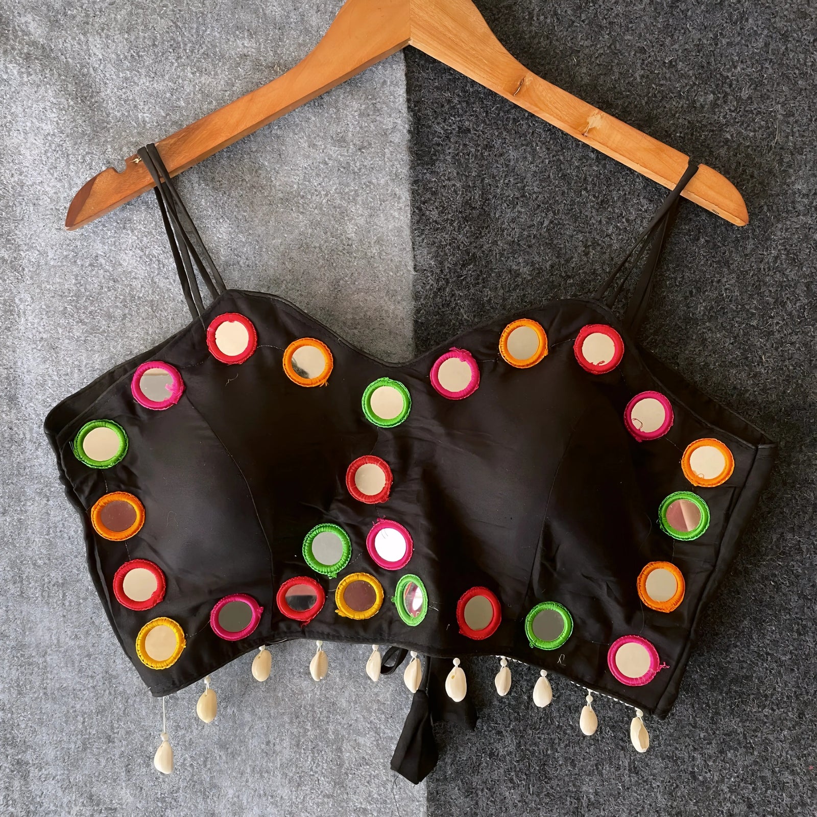 Black corset-style blouse adorned with vibrant round mirror work and multicolored embroidered borders, featuring thin shoulder straps and shell tassel drops along the hem. Displayed on a wooden hanger against a two-tone grey textured background, highlighting its festive and contemporary ethnic design.