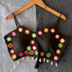 Black corset-style blouse adorned with vibrant round mirror work and multicolored embroidered borders, featuring thin shoulder straps and shell tassel drops along the hem. Displayed on a wooden hanger against a two-tone grey textured background, highlighting its festive and contemporary ethnic design.
