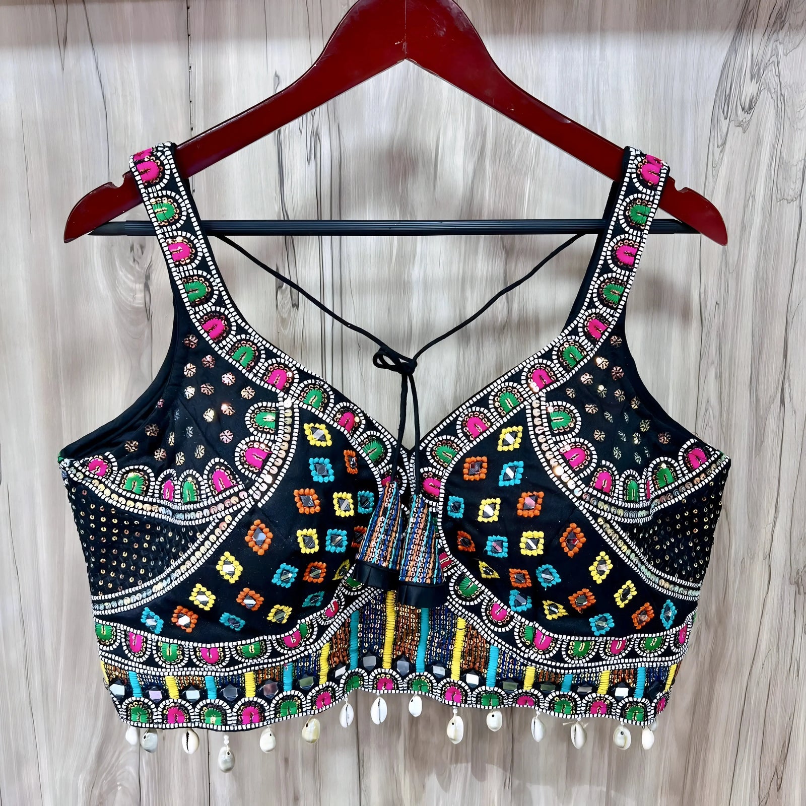 Traditional black blouse featuring vibrant multicolor Kutch-style embroidery with geometric patterns in yellow, orange, blue, green, and pink, accented with intricate beadwork, white cowrie shell trim along the hem, and decorative tie-back detail. The blouse is displayed on a wooden hanger against a light wood background.
