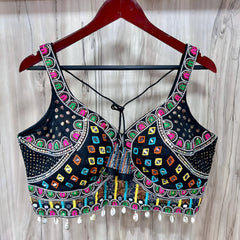 Traditional black blouse featuring vibrant multicolor Kutch-style embroidery with geometric patterns in yellow, orange, blue, green, and pink, accented with intricate beadwork, white cowrie shell trim along the hem, and decorative tie-back detail. The blouse is displayed on a wooden hanger against a light wood background.
