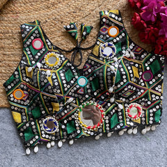 Traditional black Gujarati-style blouse richly adorned with vibrant multicolor geometric embroidery and circular mirror work, decorated with colorful thread rings, white cowrie shell trim along the hem, and tasseled tie-back detail, displayed flat on a grey carpet with a woven mat and fuchsia artificial flowers beside it.