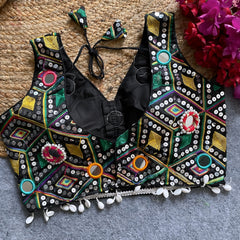 Traditional black Gujarati-style blouse richly adorned with vibrant multicolor geometric embroidery and circular mirror work, decorated with colorful thread rings, white cowrie shell trim along the hem, and tasseled tie-back detail, displayed flat on a grey carpet with a woven mat and fuchsia artificial flowers beside it.