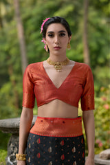A woman models a striking Black Banarasi Silk Saree with scattered red and gold Zari motifs. The saree is framed by a wide, opulent Terracotta Red border and pallu featuring dense traditional gold weaving. She wears a red blouse and heavy gold Kundan jewelry, posing outdoors on a balcony.