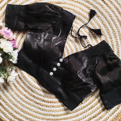 Black satin blouse with pleated detailing and three decorative silver buttons on the front, featuring elbow-length sleeves and tie-up tassels, displayed on a textured circular mat with pink and white flowers on the side.