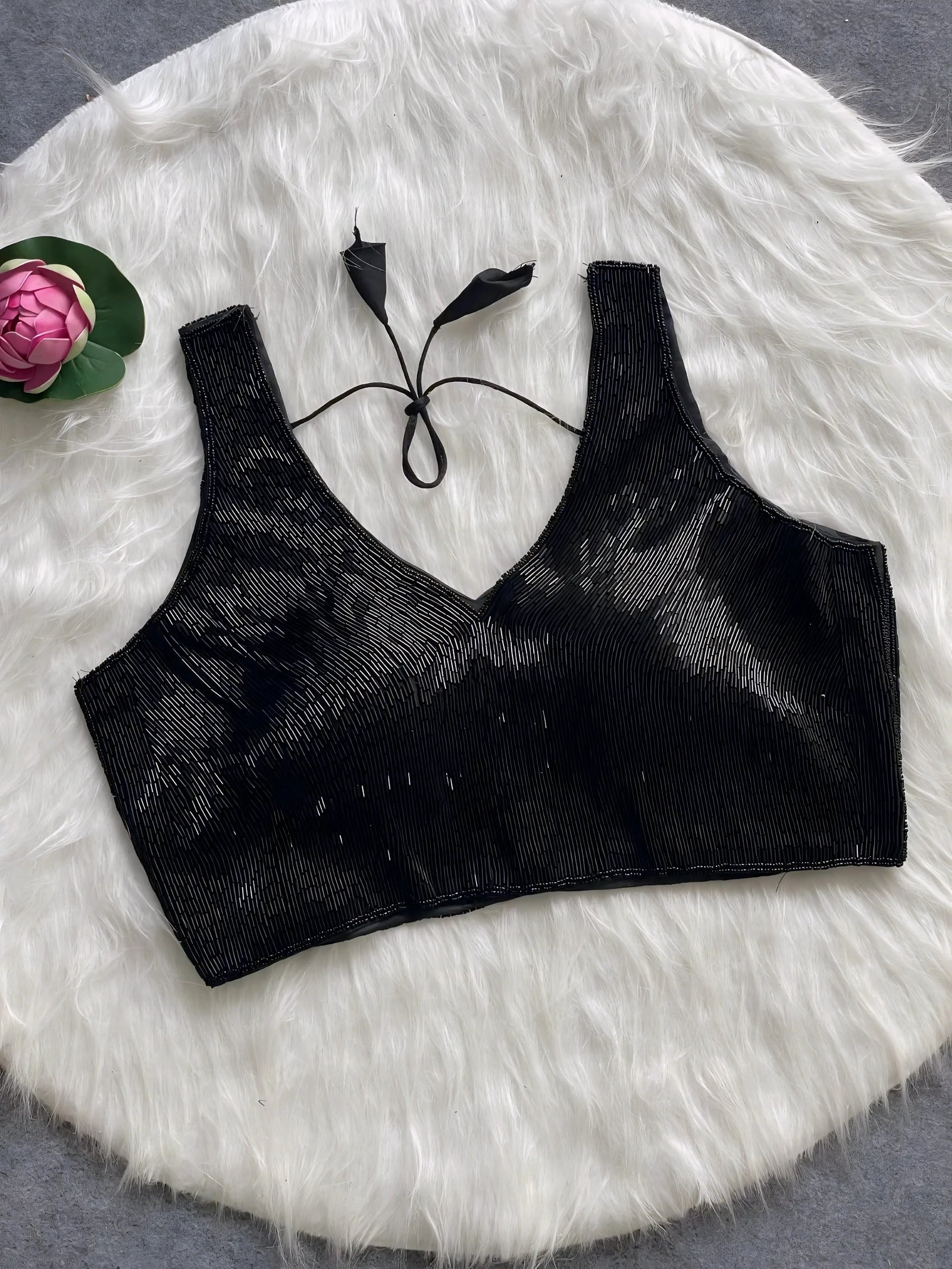 Black sequin sleeveless blouse with tie-back detail, displayed on a white fur background with a pink floral prop in the corner.