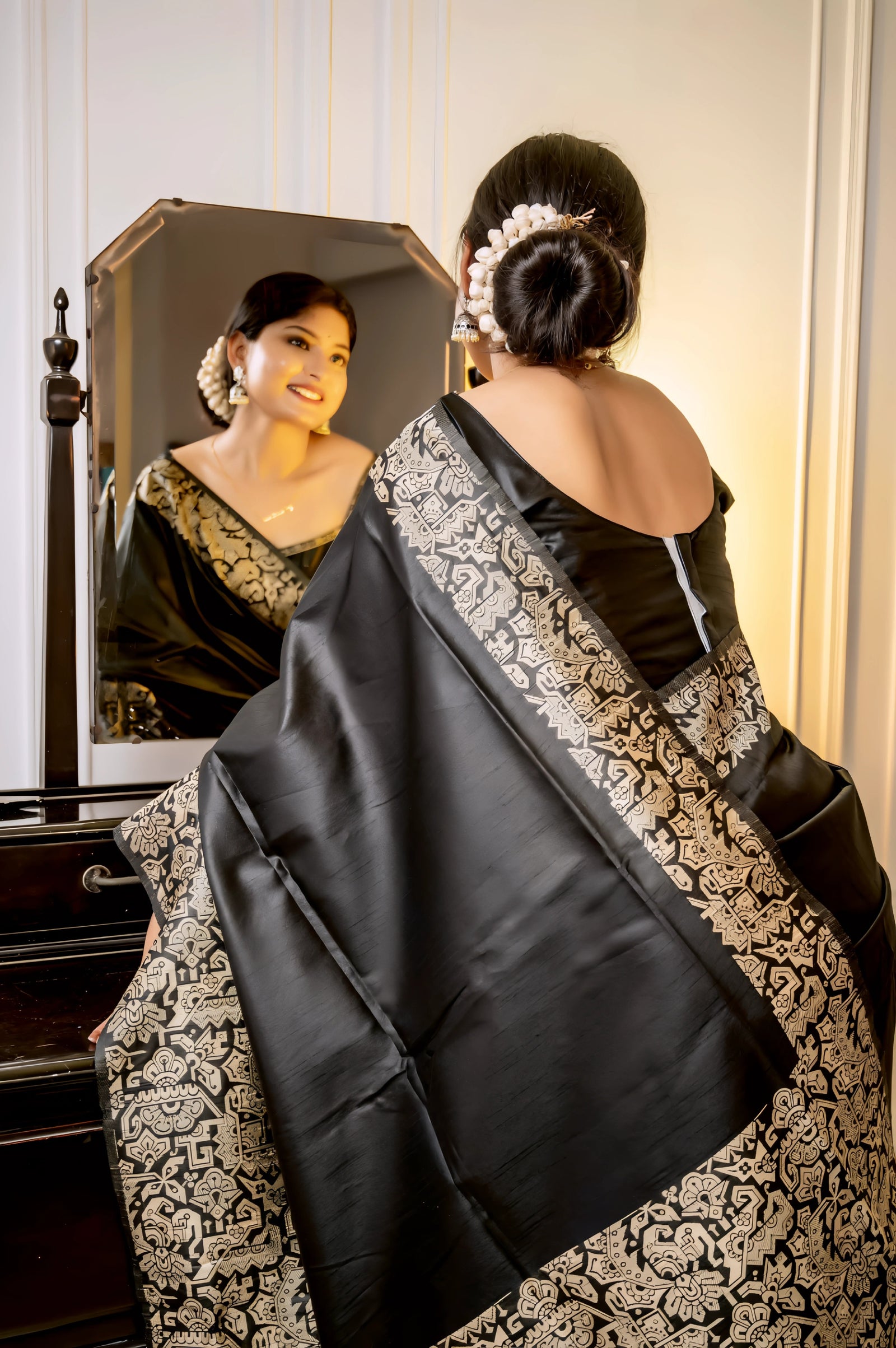 A woman models a luxurious Deep Black Raw Silk Saree. The saree features a wide border and pallu with dense, intricate gold Zari weaving in a Kalamkari-style floral and paisley pattern. She wears a matching short-sleeve black blouse and elegant gold jewelry, with white flowers adorning her hair, posing indoors next to a dark wooden vanity.