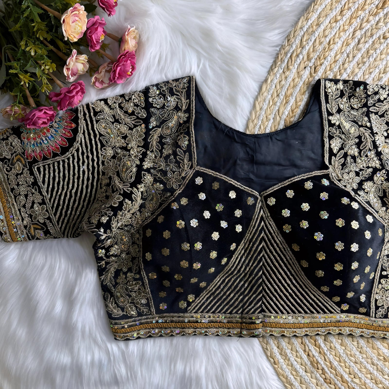 Traditional black silk blouse featuring intricate gold zardosi embroidery with floral motifs, sequin polka dot patterns, geometric pinstripes, and a colorful peacock feather motif on the sleeve, displayed flat on a white fur surface with a braided mat and artificial pink flowers in the corner.