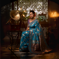 A woman models a luxurious Aqua Blue Soft Banarasi Silk Saree. The saree features heavy dual-toned gold and dark Zari weaving with a prominent dark brown/maroon accent border. She wears a contrasting mauve short-sleeve blouse and a heavy gold and stone choker necklace, posing in a dramatically lit vintage setting