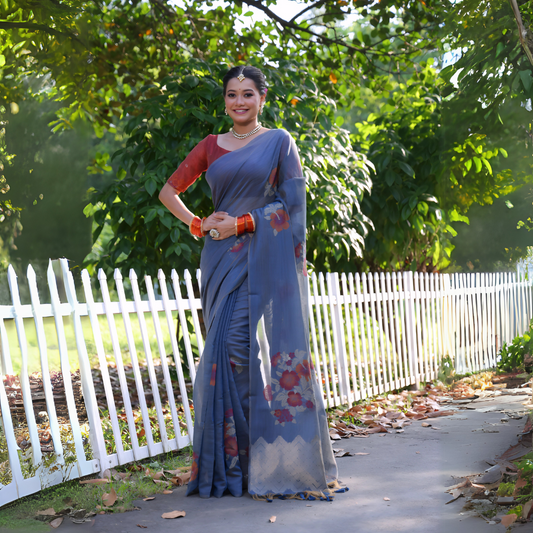 A woman models an elegant Slate Blue Soft Muga Cotton Saree. The pallu features large, colorful Resham floral weaving in red and orange. She wears a contrasting terracotta red blouse and is accessorized with a pearl choker necklace and vibrant orange bangles, posing outdoors by a white fence.