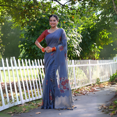 A woman models an elegant Slate Blue Soft Muga Cotton Saree. The pallu features large, colorful Resham floral weaving in red and orange. She wears a contrasting terracotta red blouse and is accessorized with a pearl choker necklace and vibrant orange bangles, posing outdoors by a white fence.
