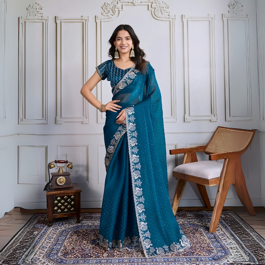 Woman wearing a teal blue georgette saree embellished with sparkly sequins and an ornate silver embroidered floral border, paired with a matching blouse with delicate embroidery. She stands elegantly in a classic white paneled room, accessorized with statement jhumka earrings and bangles, next to a vintage rotary telephone on a carved wooden side table and a modern wooden armchair, all atop a richly patterned rug for a sophisticated, timeless look.
