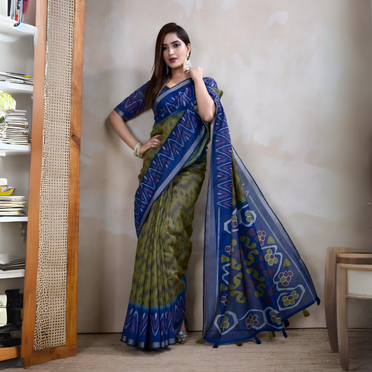 Woman wearing a blue and green Ikat silk saree featuring a geometric zigzag pattern and a striking blue pallu with floral and abstract motifs, paired with a matching blouse and traditional silver jewelry. She stands gracefully in a contemporary studio with a textured neutral wall, a woven cane screen, wooden furniture, and a bookshelf, creating an elegant and modern aesthetic.