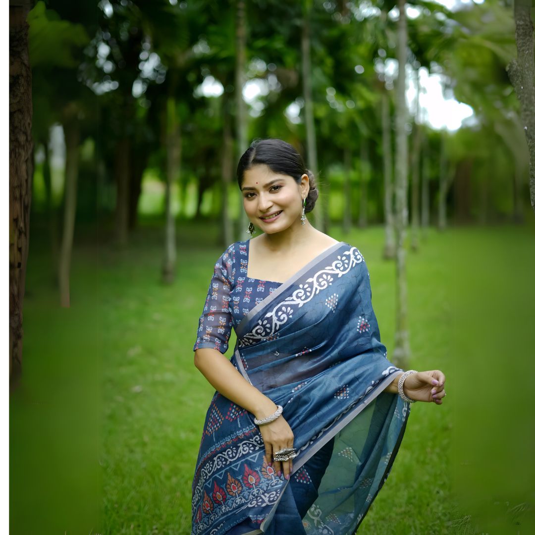 A woman models a deep Indigo Blue Soft Cotton Saree featuring various digital block prints, including geometric and colorful traditional motifs. She wears a printed blue blouse and large oxidized silver jewelry, posing in a lush green outdoor setting.