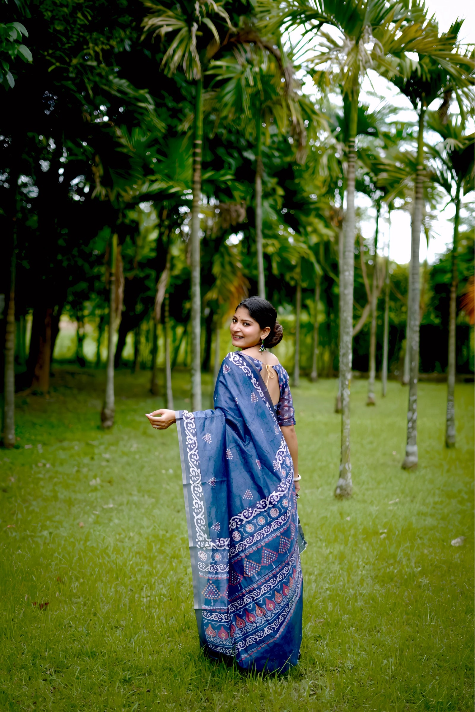 A woman models a deep Indigo Blue Soft Cotton Saree featuring various digital block prints, including geometric and colorful traditional motifs. She wears a printed blue blouse and large oxidized silver jewelry, posing in a lush green outdoor setting.