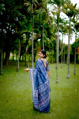 A woman models a deep Indigo Blue Soft Cotton Saree featuring various digital block prints, including geometric and colorful traditional motifs. She wears a printed blue blouse and large oxidized silver jewelry, posing in a lush green outdoor setting.