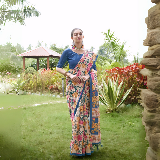 A woman models an elegant Beige Soft Tussar Silk Saree with a dense all-over print of multi-colored Kalamkari florals and vines (in pink, blue, and green). The saree has a wide, contrasting Deep Blue border and pallu with traditional print. She wears a matching blue blouse and a chunky beaded necklace, posing outdoors in a bright garden setting.