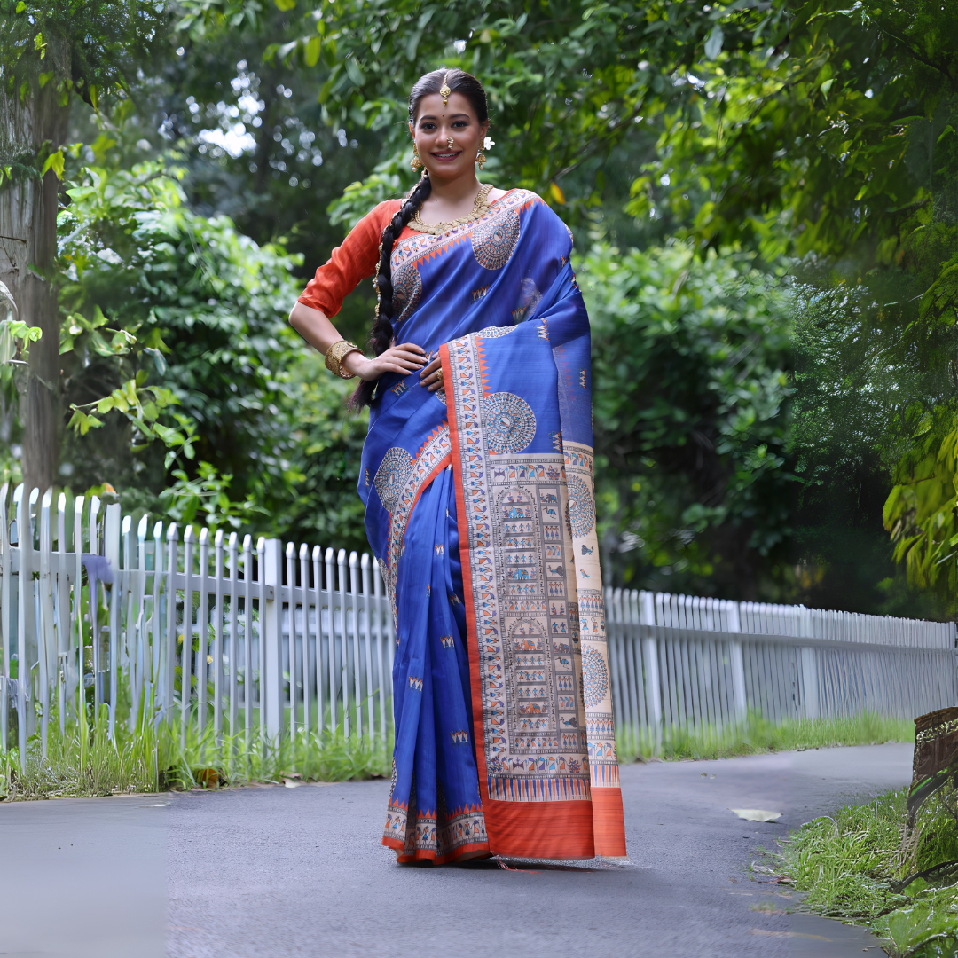 A woman models a vibrant Royal Blue Soft Tussar Silk Saree with a bright orange border. The pallu features a wide, elaborate panel with intricate Madhubani-style folk art prints in cream, blue, and orange. She wears an orange blouse and a gold choker necklace, posing outdoors.