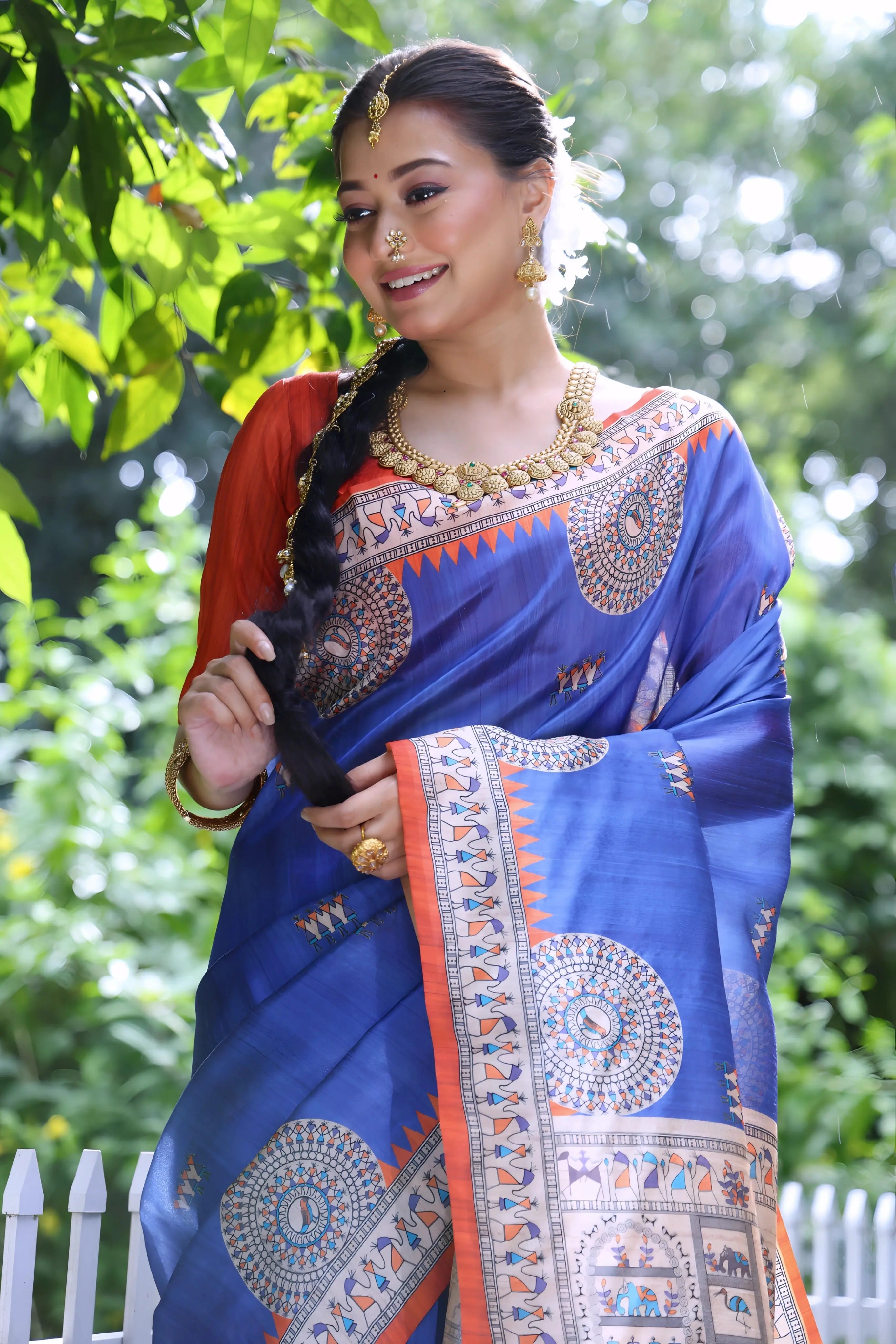 A woman models a vibrant Royal Blue Soft Tussar Silk Saree with a bright orange border. The pallu features a wide, elaborate panel with intricate Madhubani-style folk art prints in cream, blue, and orange. She wears an orange blouse and a gold choker necklace, posing outdoors.