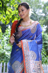 A woman models a vibrant Royal Blue Soft Tussar Silk Saree with a bright orange border. The pallu features a wide, elaborate panel with intricate Madhubani-style folk art prints in cream, blue, and orange. She wears an orange blouse and a gold choker necklace, posing outdoors.