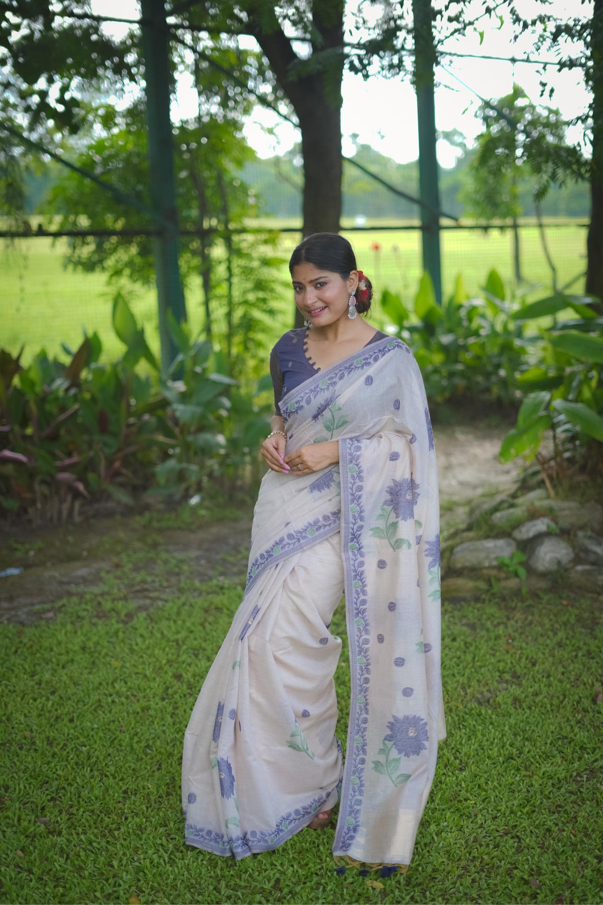 Woman in blue resham weaving saree with floral design posing gracefully in a green garden setting