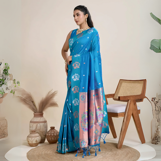 A woman models a luxurious Deep Blue Soft Silk Saree. The saree body features subtle metallic motifs. The pallu features a striking contrast panel in metallic rose gold/copper, richly woven with intricate blue and silver peacock and floral motifs. She wears a matching blue sleeveless blouse and heavy gold Kundan jewelry.