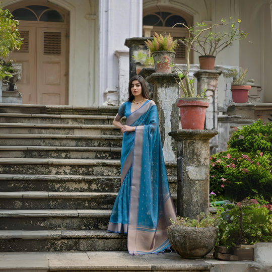 A woman models an elegant Deep Teal Soft Silk Saree. The saree features subtle metallic motifs on the body and a wide, contrasting matte silver/gray Zari woven border. She wears a dark grey short-sleeve blouse and a delicate gold choker necklace, posing gracefully on an ornate stone staircase.