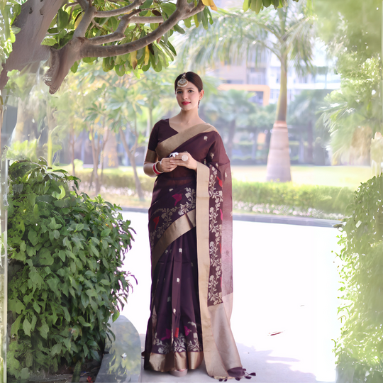A woman models an elegant Deep Plum Linen Silk Saree. The saree features delicate woven floral and bird motifs and a wide, contrasting pale gold Zari border. She wears a matching plum blouse, contrasting red and white bangles, and a traditional gold maang tikka, posing outdoors.