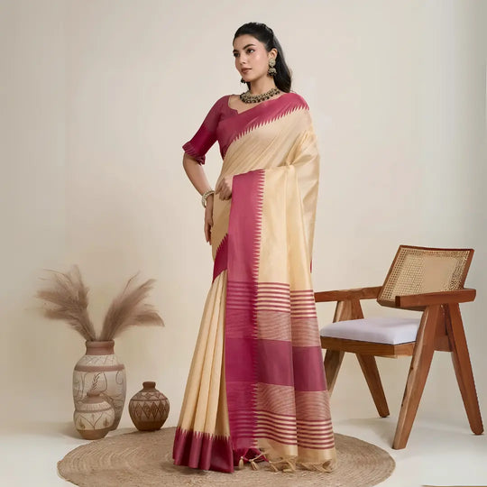 A woman models a striking Beige Raw Silk Saree. The saree has a wide, vibrant deep pink border featuring traditional temple weaving. The pallu has wide horizontal stripes in pink and gold. She wears a pink blouse and a gold and green Kundan necklace.