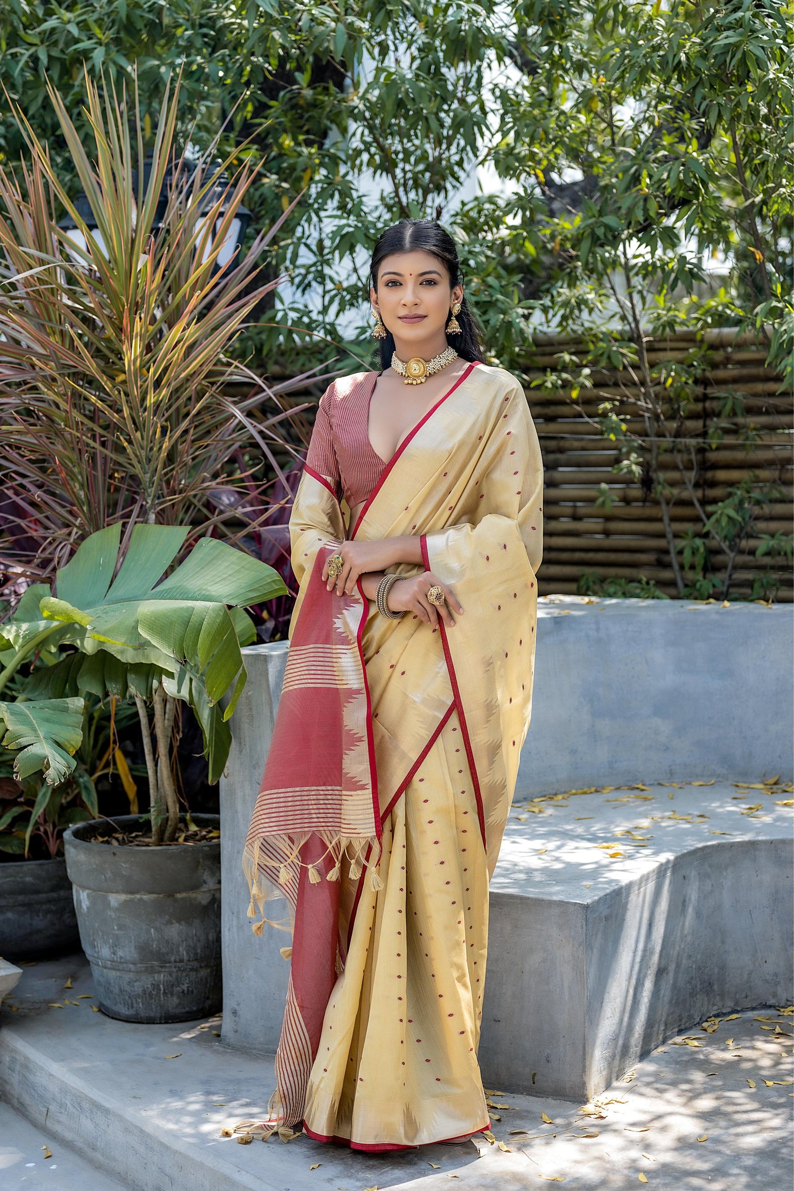 A woman models an elegant Beige Tussar Silk Saree with scattered red woven motifs. The saree has a gold border and a pallu featuring wide bands of deep red and gold Zari weaving. She wears a red blouse and heavy gold Kundan jewelry, posing outdoors.