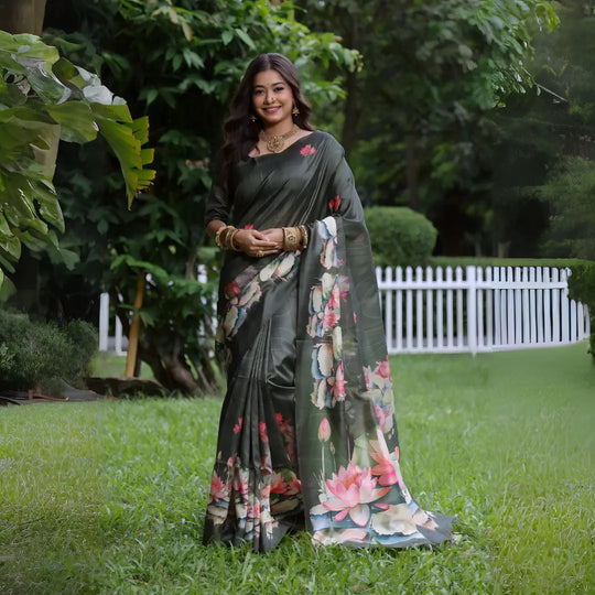 A woman models a stunning Dark Olive Green Soft Tussar Silk Saree. The saree features a large-scale digital print of colorful floral and lotus motifs in pink, white, and pale blue on the pallu and lower drape. She wears a matching elbow-sleeve green blouse and heavy gold Kundan jewelry, posing outdoors in a garden setting.