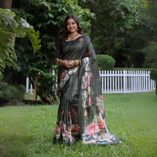A woman models a stunning Dark Olive Green Soft Tussar Silk Saree. The saree features a large-scale digital print of colorful floral and lotus motifs in pink, white, and pale blue on the pallu and lower drape. She wears a matching elbow-sleeve green blouse and heavy gold Kundan jewelry, posing outdoors in a garden setting.