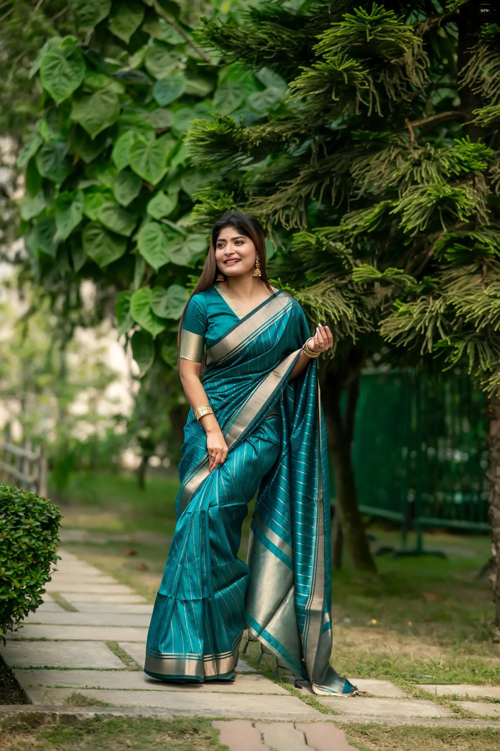 Woman in a dark green saree with zari work standing outdoors amidst lush greenery