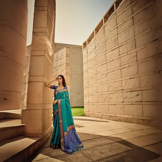 Graceful woman showcasing deep green saree with intricate zari work and detailed pallu, standing by modern sandstone structures under sunny sky