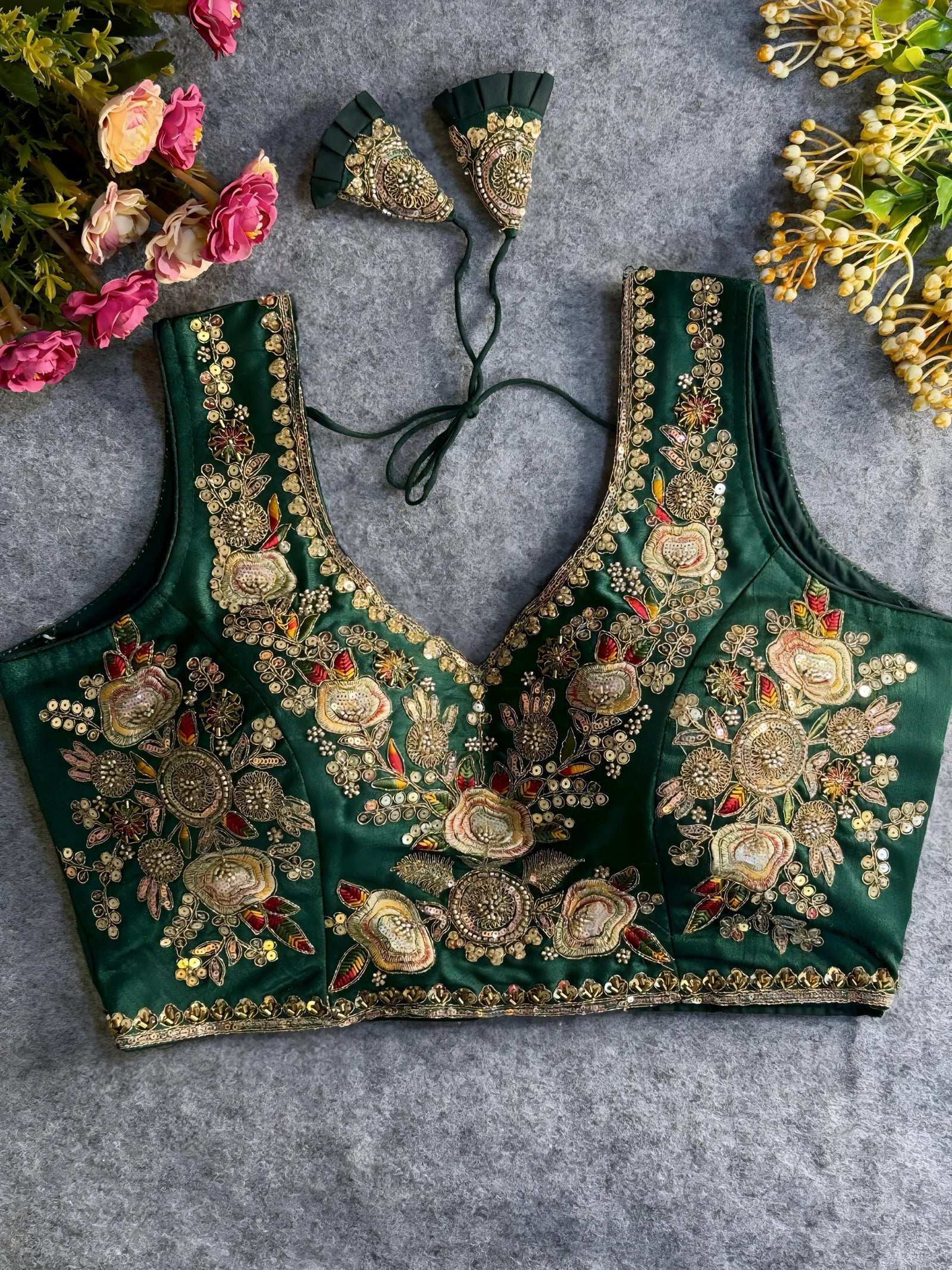 Traditional emerald green silk blouse featuring intricate gold zardozi embroidery and beadwork with multicolor floral and leaf motifs on the front and neckline, finished with decorative tassel ties, displayed flat on a textured grey background with artificial pink and yellow flowers in the corners.