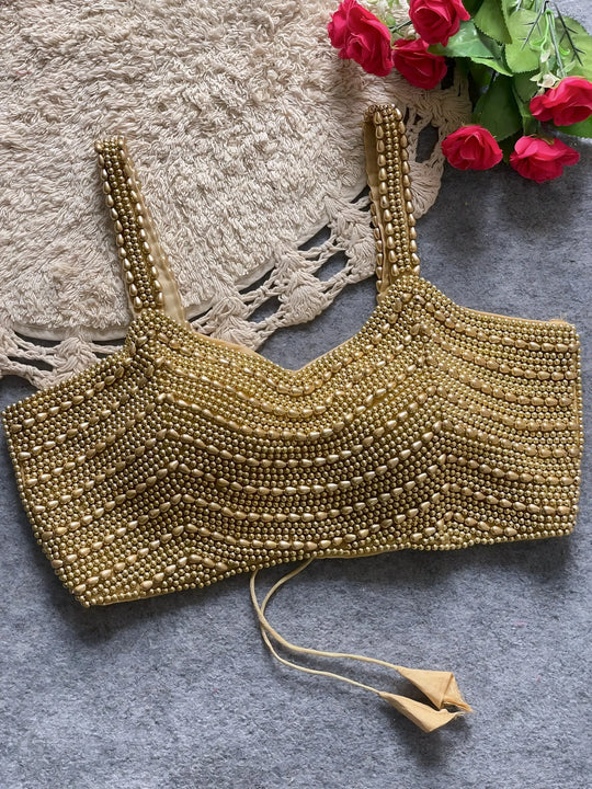 Traditional gold blouse featuring intricate beaded and sequin embellishments in horizontal rows, wide shoulder straps, and a tasseled tie-back detail, displayed flat on a textured grey surface with a braided cream mat and red artificial roses in the corner.