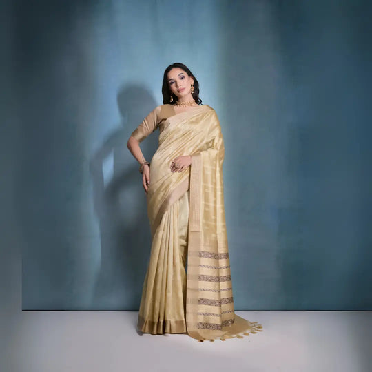 A woman models an elegant Beige/Champagne Soft Raw Silk Saree with a wide metallic gold Zari border. The pallu features distinct horizontal bands of dark brown/black Ikat-style geometric weaving, providing a strong contrast. She wears a matching beige blouse and accessorizes with a gold choker necklace and large gold earrings, posing indoors.