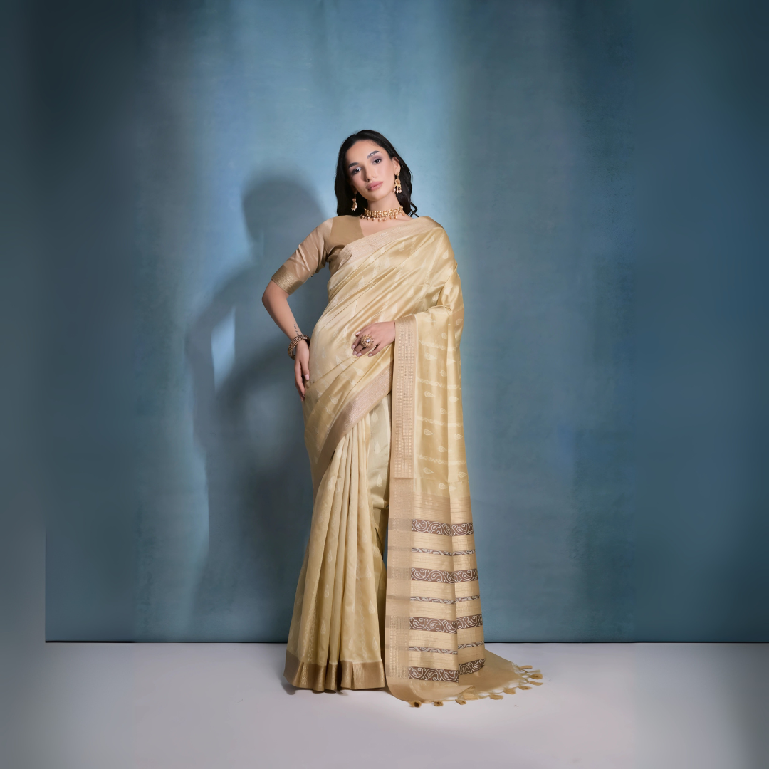 A woman models an elegant Beige/Champagne Soft Raw Silk Saree with a wide metallic gold Zari border. The pallu features distinct horizontal bands of dark brown/black Ikat-style geometric weaving, providing a strong contrast. She wears a matching beige blouse and accessorizes with a gold choker necklace and large gold earrings, posing indoors.