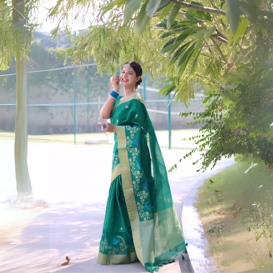 A woman models a vibrant Emerald Green Linen Silk Saree with delicate Zari woven floral and bird motifs. The saree has a wide, contrasting pale gold/pistachio green border. She wears bold blue bangles and a headpiece, posing outdoors.