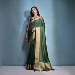 A woman models a striking Dark Green Banarasi Raw Silk Saree. The saree body features subtle gold Zari stripes and is framed by an extremely wide, luxurious metallic gold Zari border. She wears a sleeveless dark blouse and traditional gold Kundan jewelry, posing indoors.