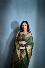 A woman models a striking Dark Green Banarasi Raw Silk Saree. The saree body features subtle gold Zari stripes and is framed by an extremely wide, luxurious metallic gold Zari border. She wears a sleeveless dark blouse and traditional gold Kundan jewelry, posing indoors.