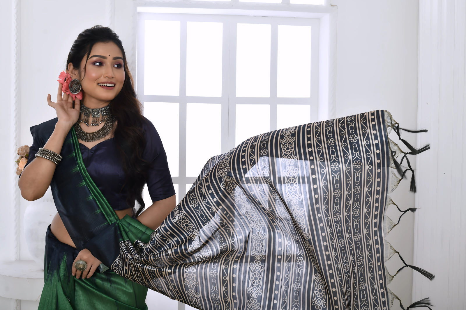 A woman models a dramatic Two-Toned Green Soft Tussar Silk Saree. The saree features bold black geometric patterns along the border and a pallu with dense abstract black, grey, and white tribal-style print. She wears a black blouse and chunky oxidized silver jewelry, posing indoors by a window.
