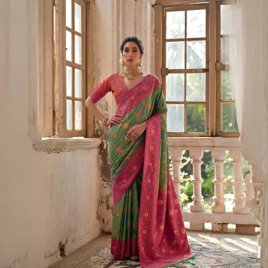 A woman models a striking Moss Green Banarasi Silk Saree with gold Zari weaving. The saree has a wide, ornate Fuchsia Pink border and pallu featuring traditional geometric Zari weaving. She wears a pink and gold blouse and heavy gold Kundan jewelry.