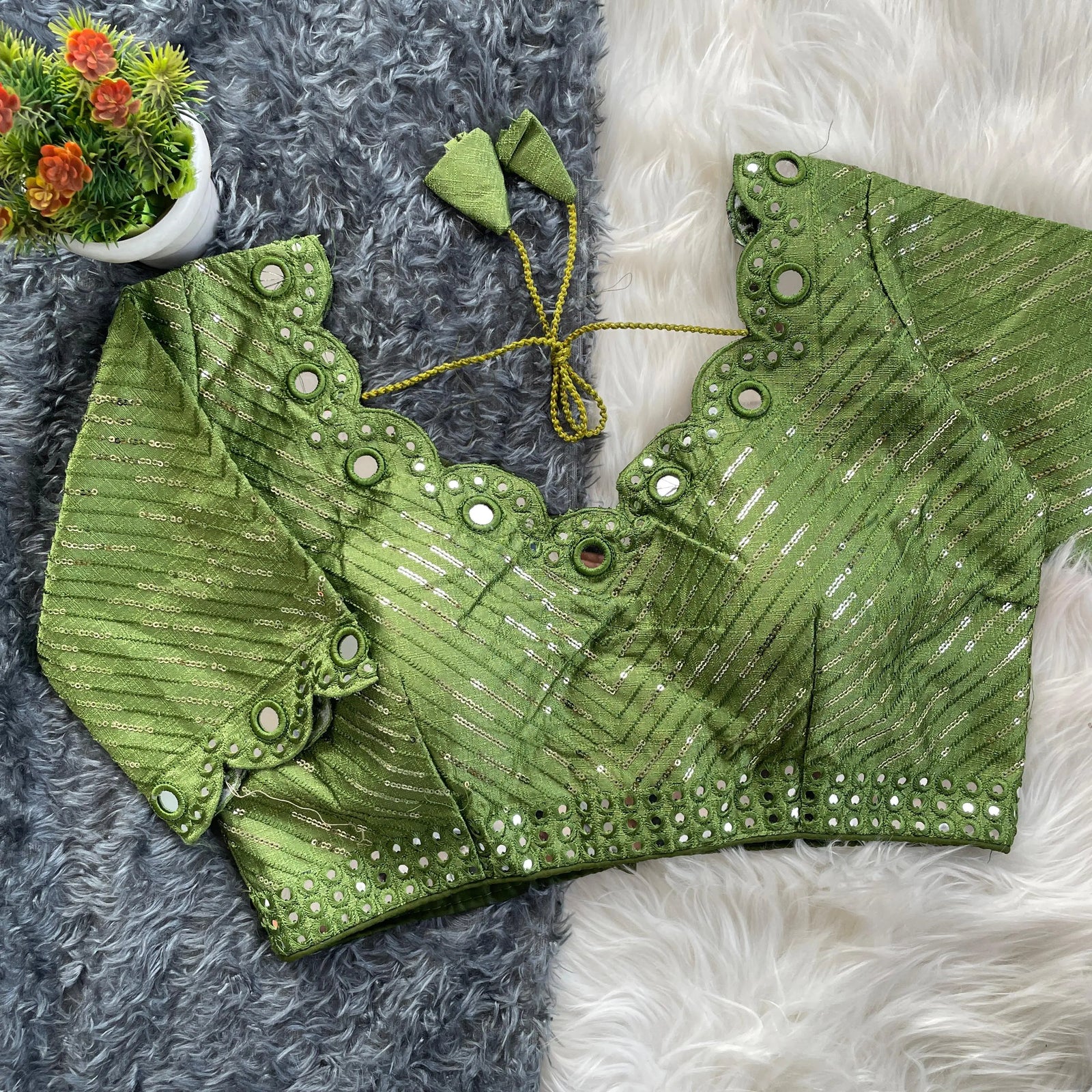 Green bridal blouse with scallop sleeves, featuring silver sequin stripes and mirror work detailing along the neckline, sleeves, and hem, displayed on a textured fur background with a small potted plant nearby.