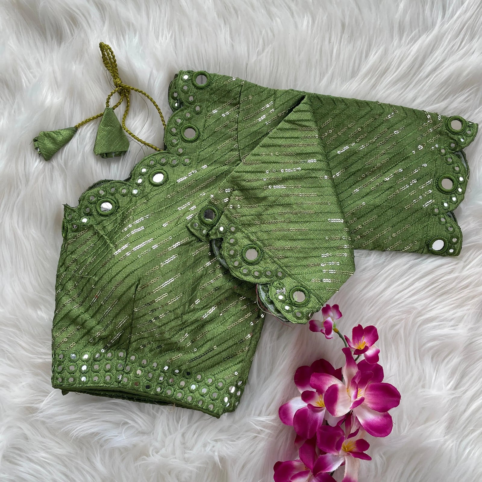 Green bridal blouse with scallop sleeves, featuring silver sequin stripes and mirror work detailing along the neckline, sleeves, and hem, displayed on a textured fur background with a small potted plant nearby.