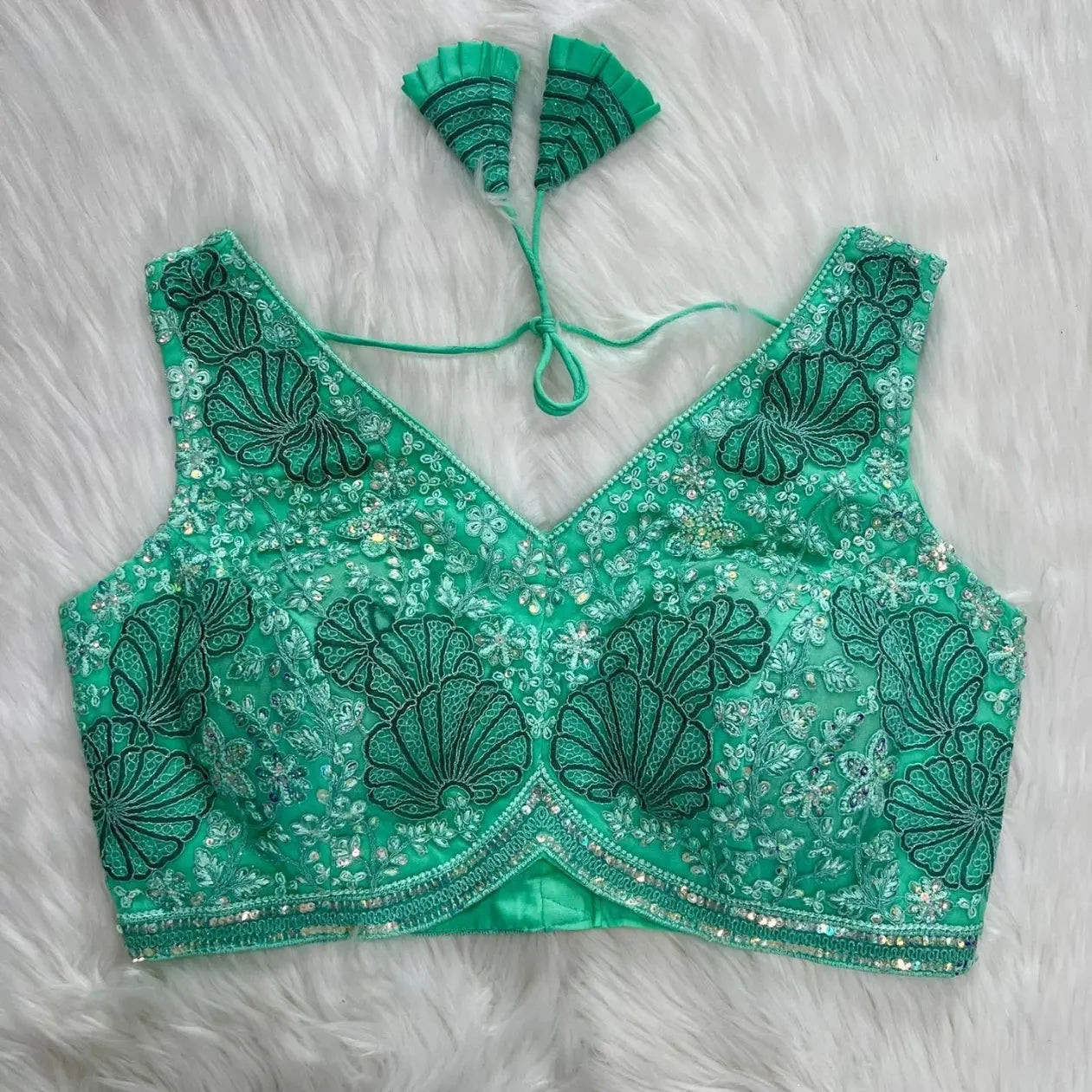 Aqua green blouse featuring intricate shell-shaped embroidery, sparkling sequin and bead embellishments, floral motifs, and a geometric border, with a deep V-neck, structured cups, and pleated fan-shaped tasseled tie-back detail, displayed flat on a white fur background.
