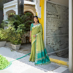 A woman models a vibrant Pista Green Tussar Silk Saree with small woven motifs. The saree has a contrasting peacock blue/teal pallu featuring wide gold Zari stripes. She wears a teal blouse and gold Kundan jewelry, posing in an outdoor courtyard.