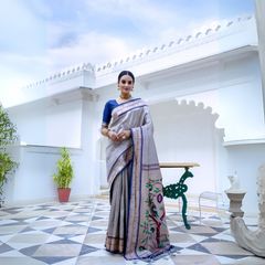 A woman models an elegant Light Gray Soft Silk Saree. The saree features a striking deep navy blue contrast border accented with gold Zari. The pallu is a focal point, featuring rich, artistic Paithani-style print or weaving of colorful bird-on-branch motifs. She wears a navy blue blouse and traditional gold jewelry with green accents, posing on an ornate white terrace.